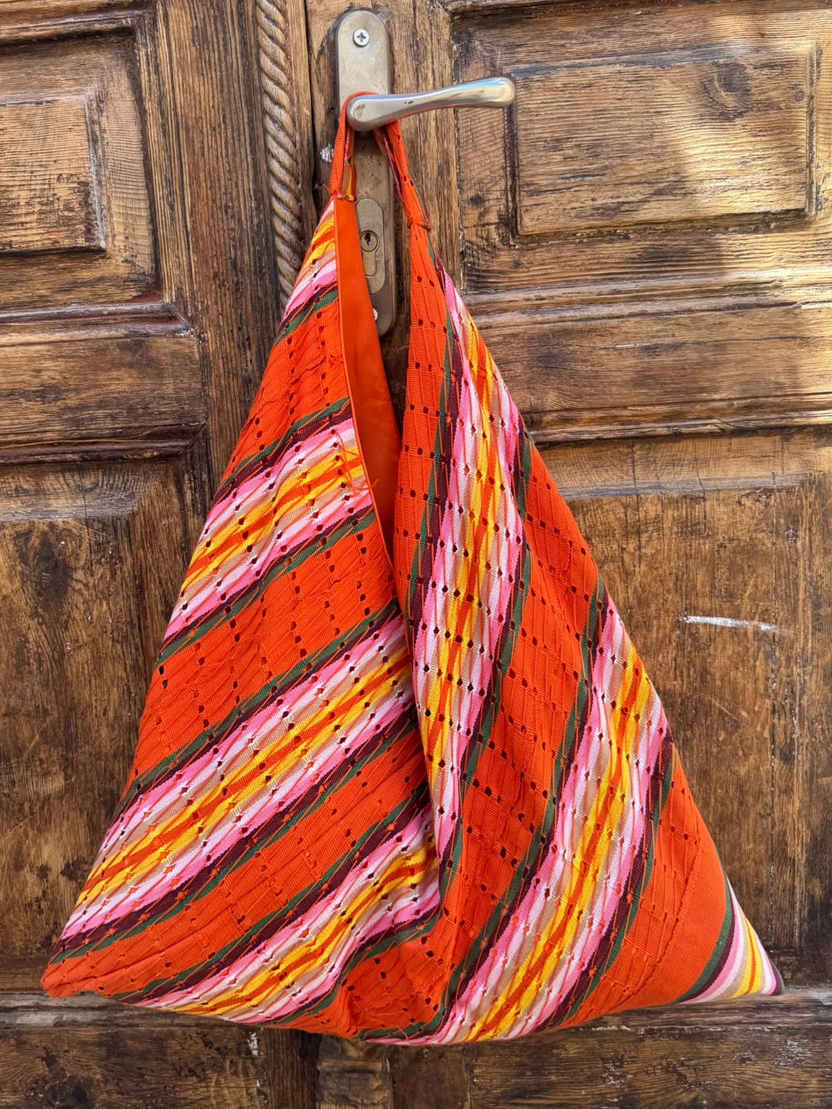 A handmade triangular shoulder bag made from orange as oke fabric with pink, yellow, and white striped kantha stitching, hanging from a metal hook on a rustic wooden door.